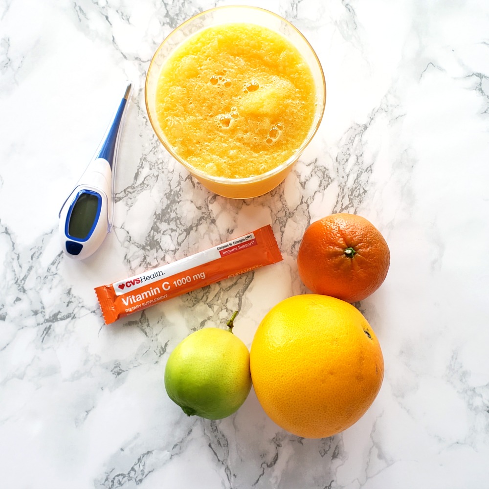 Sore Throat Slushy in a clear glass along with a tangerine, orange, lime, packet of Vitamin C and thermometer on a white marble countertop. 
