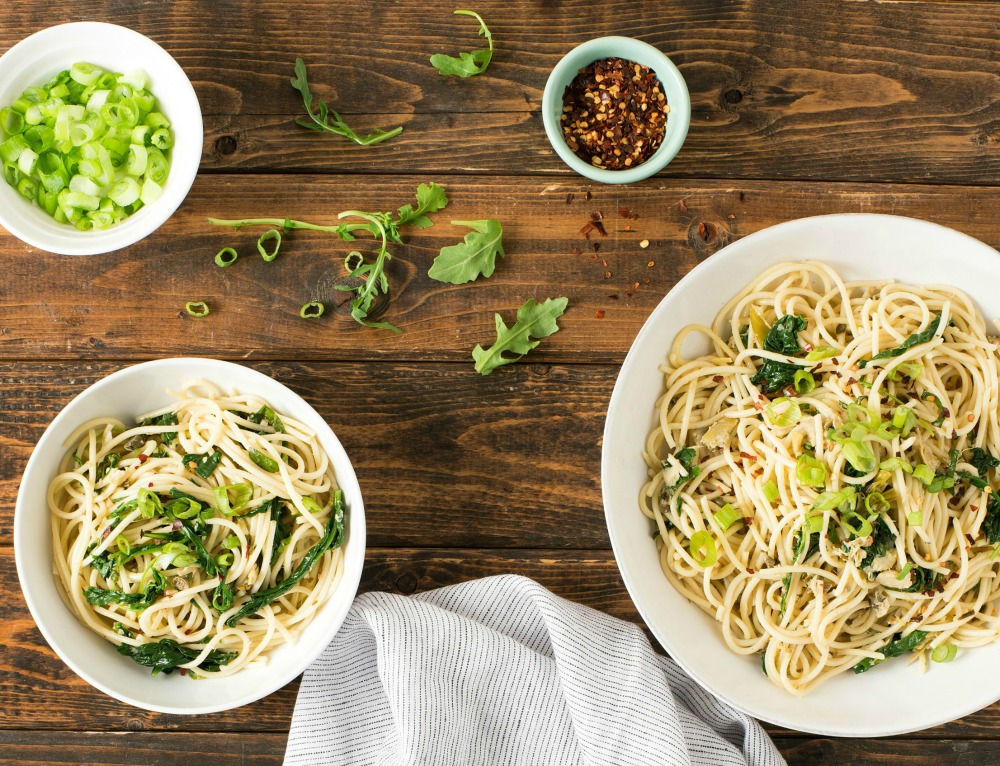 Green Pasta Puttanesca in two white bowls, chopped green onions and crushed red pepper in other bowls, on a wooden table