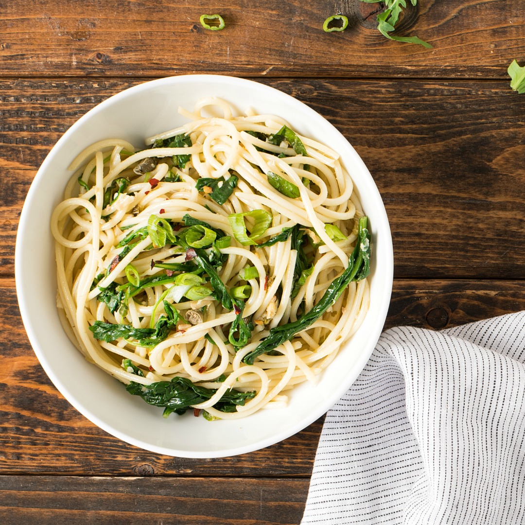 Green Pasta Puttanesca in a white bowl on a wooden table with striped towel in the corner