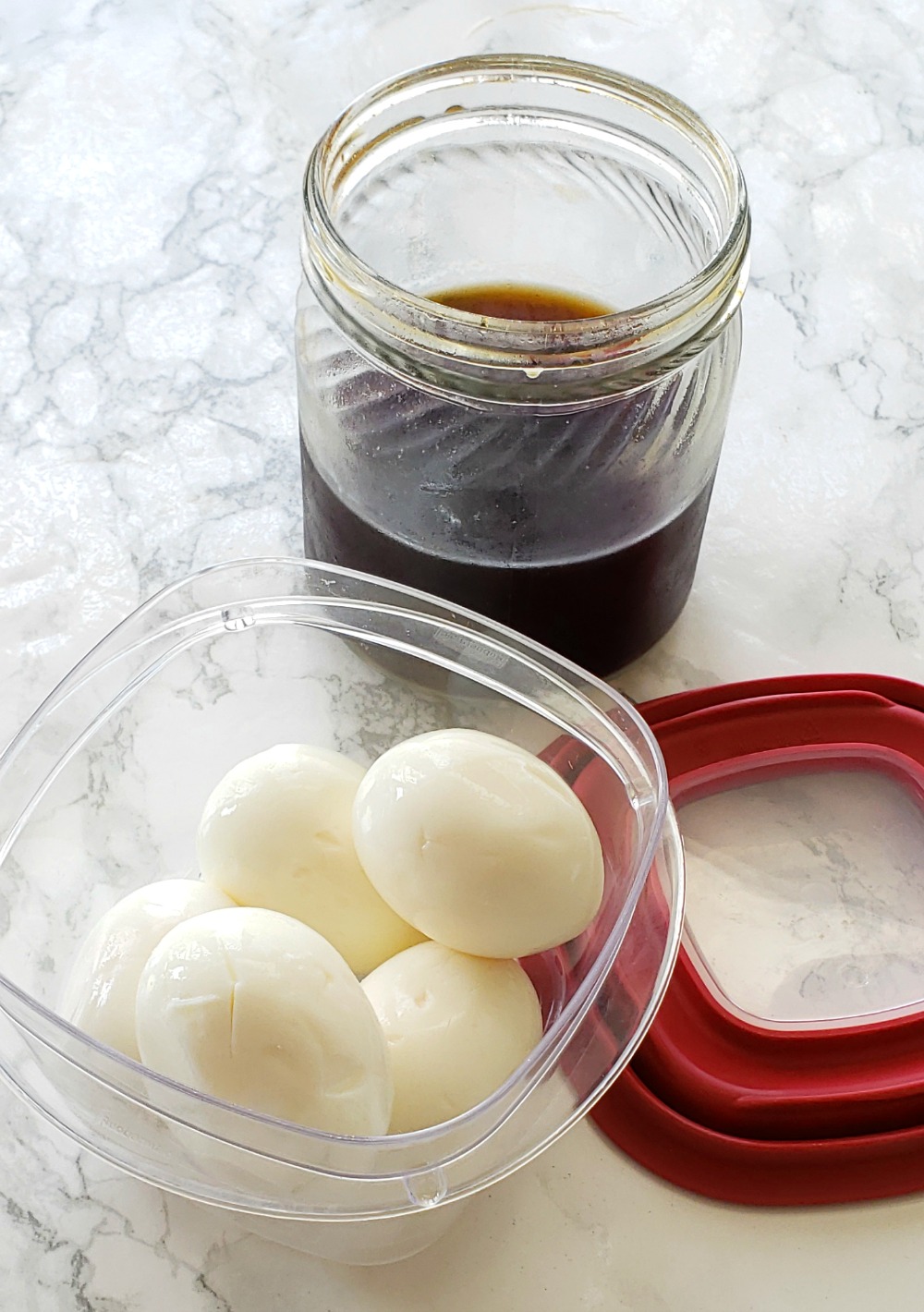 Clear container of 5 hard-boiled eggs sits on a white marble countertop with red rimmed lid alongside and dark liquid in a jar in the background