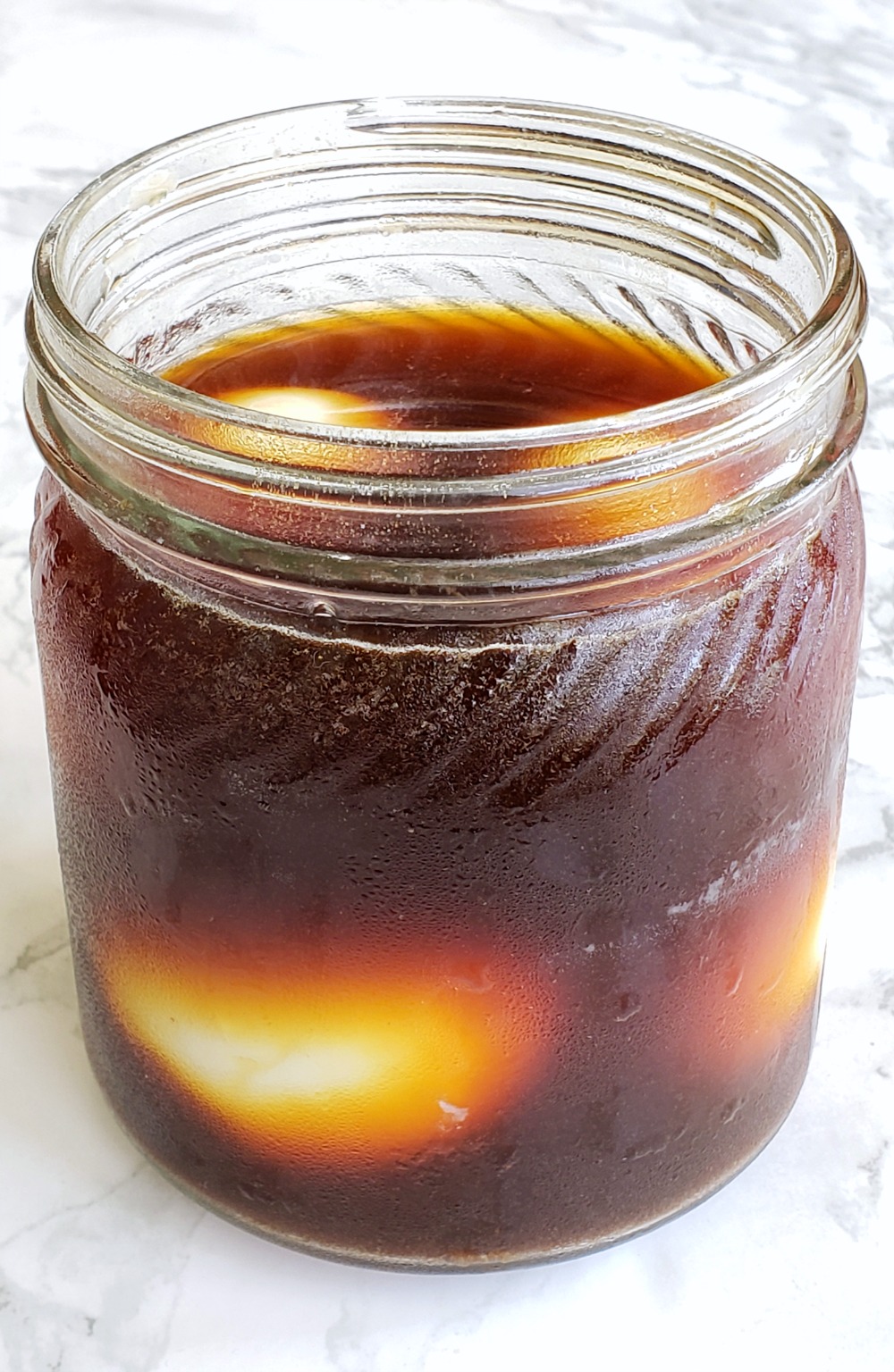 Hard-Boiled Eggs in soy brine in a clear glass jar on a white marble countertop