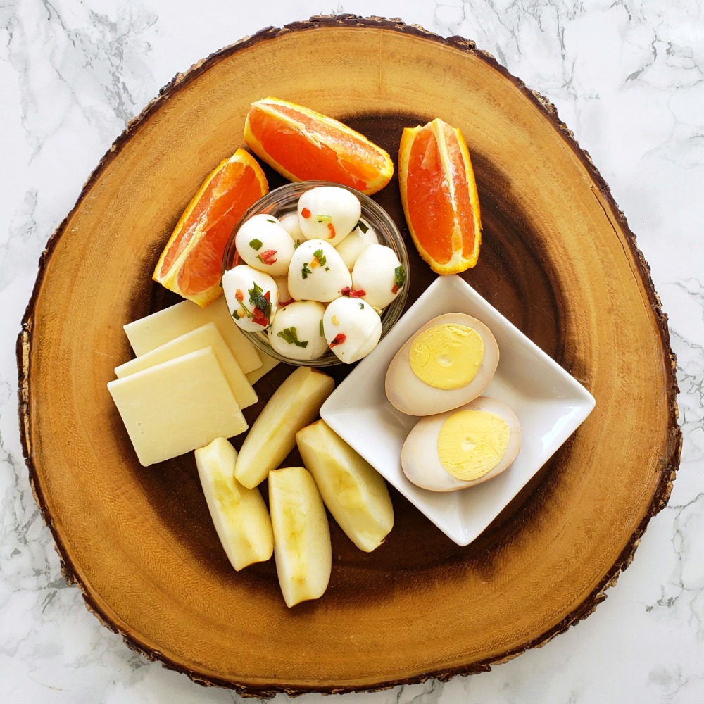 Cut cheese squares, cut orange and apple, marinated mozzarella balls and a cut open Soy-Brined Hard-Boiled Egg are atop a round wooden platter, sitting on a white marble countertop. 