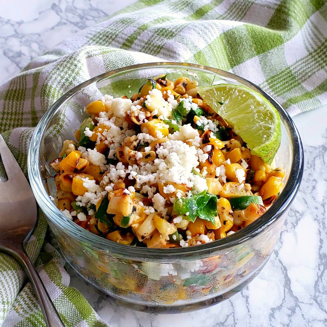 Skillet Esquites in a glass bowl with a lime slice, on a plaid green towel on a white marble counter