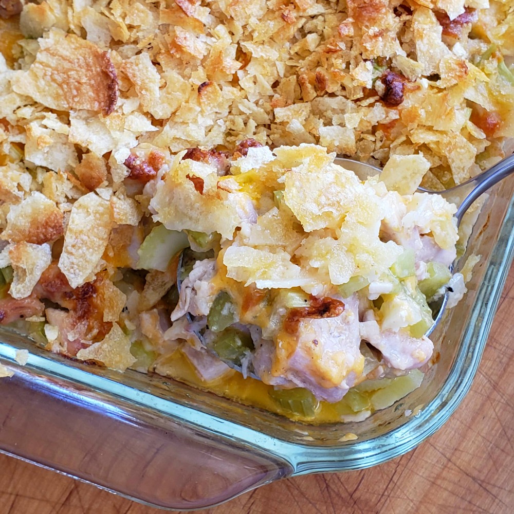 Serving spoon shows a scoop of Hot Chicken Salad Casserole in a glass baking dish on a wooden counter