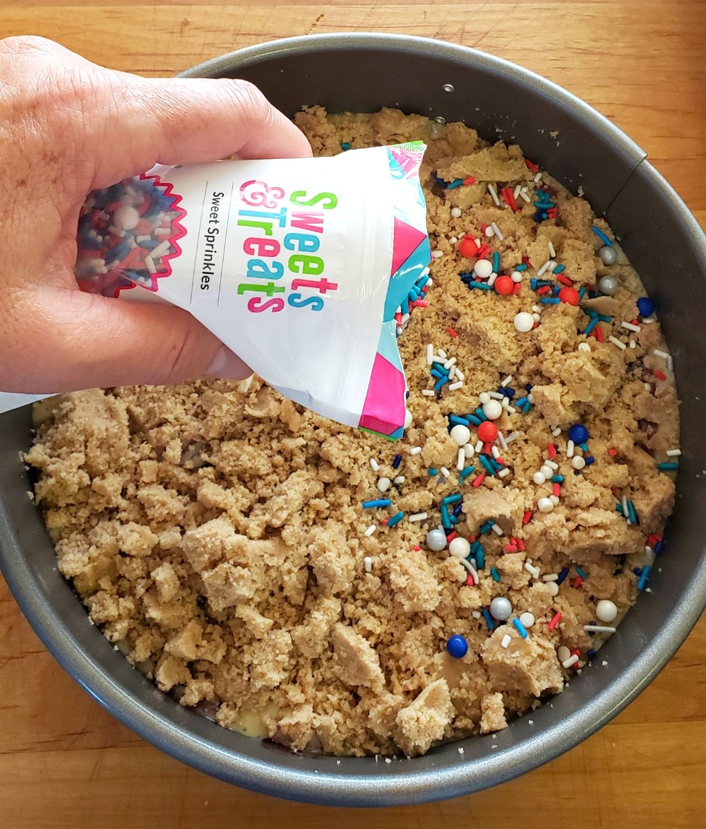 Hand holds bag of sprinkles to apply to top of Jam Breakfast Cake with Streusel on ShockinglyDelicious.com