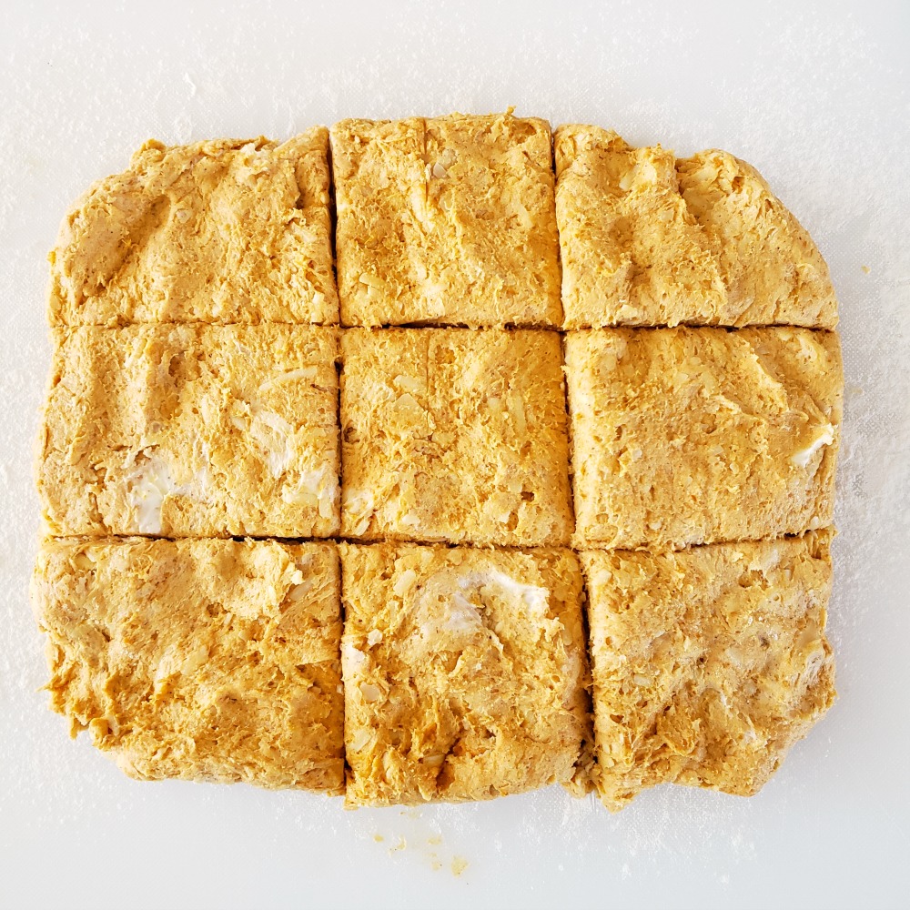 Orange pumpkin dough patted into a rectangle on a white cutting board, scored into 9 square biscuits on ShockinglyDelicious.com