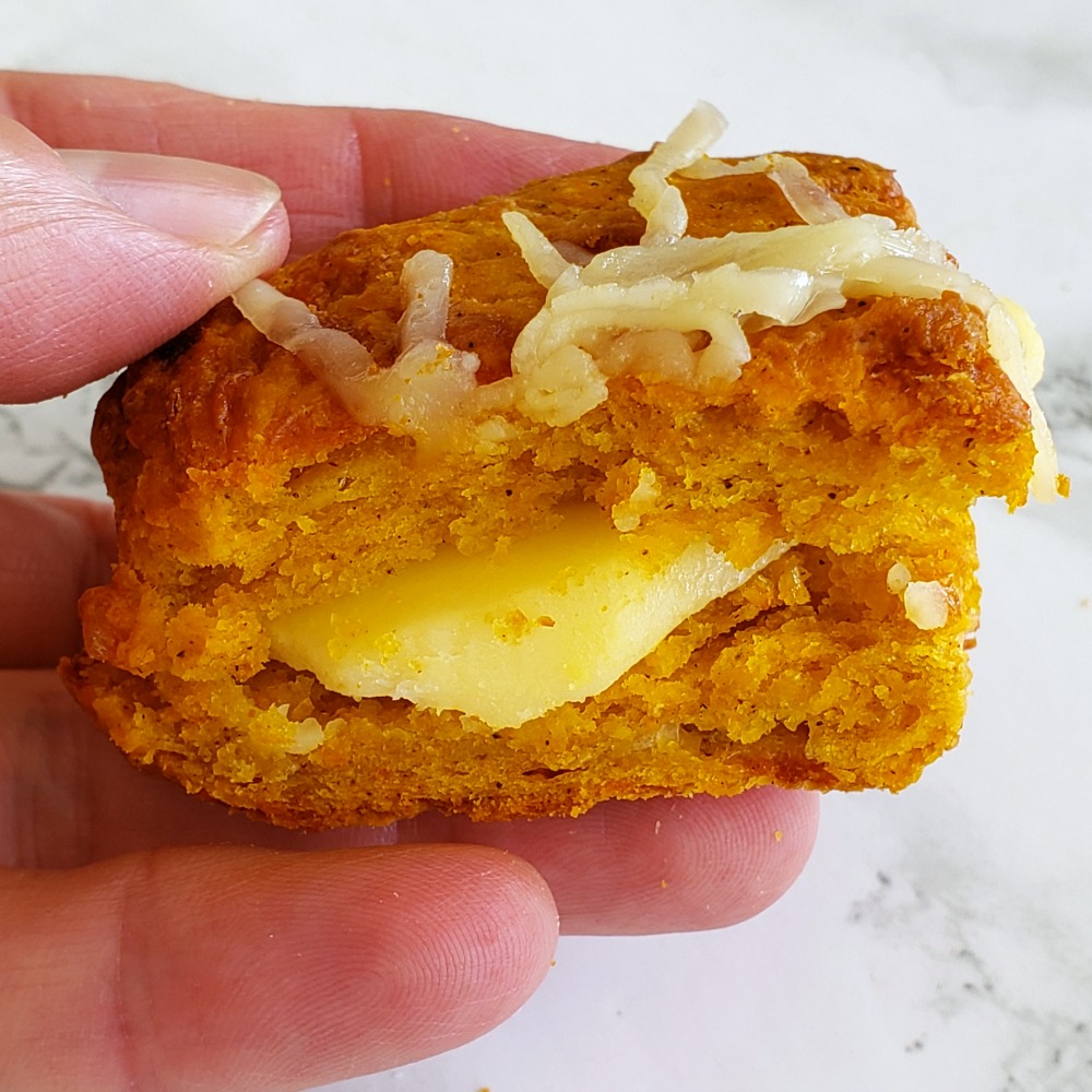 Hand holds an orange-colored Pumpkin Cheddar Biscuit stuffed with yellow cheese, with cheese shreds on top, against a white background