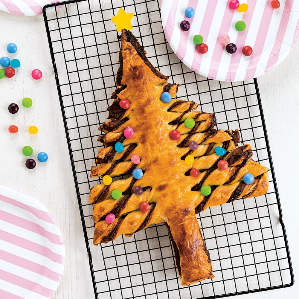 Pastry shaped like a Christmas tree with colored candy on the branches on a black wire cooling rack with pink striped paper plates on either side