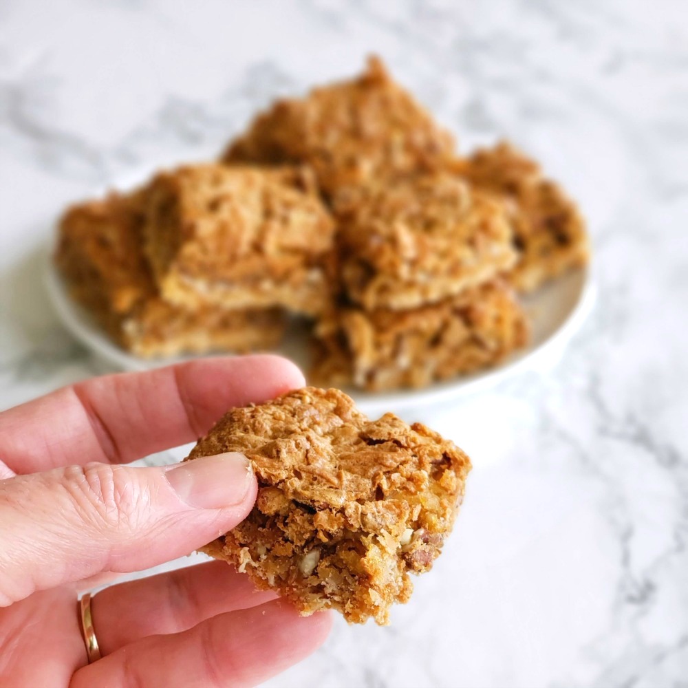 Fingers hold a single coconut pecan bar, in front of a plate of bars behind