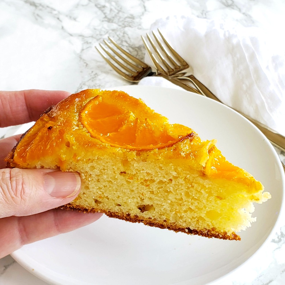 Hand holds a piece of cake over a white plate with forks in background