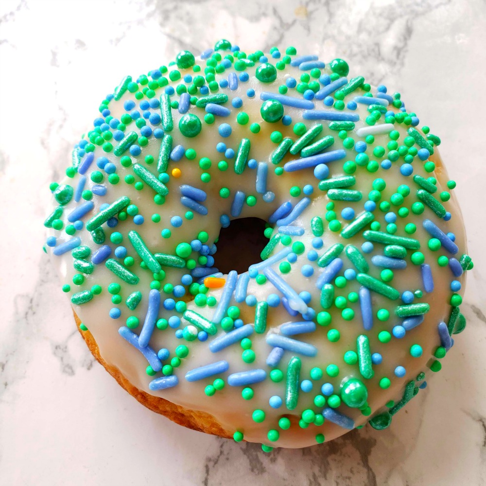 A Glazed Baked Donut with green and blue sprinkles sitting on a white marble counter.