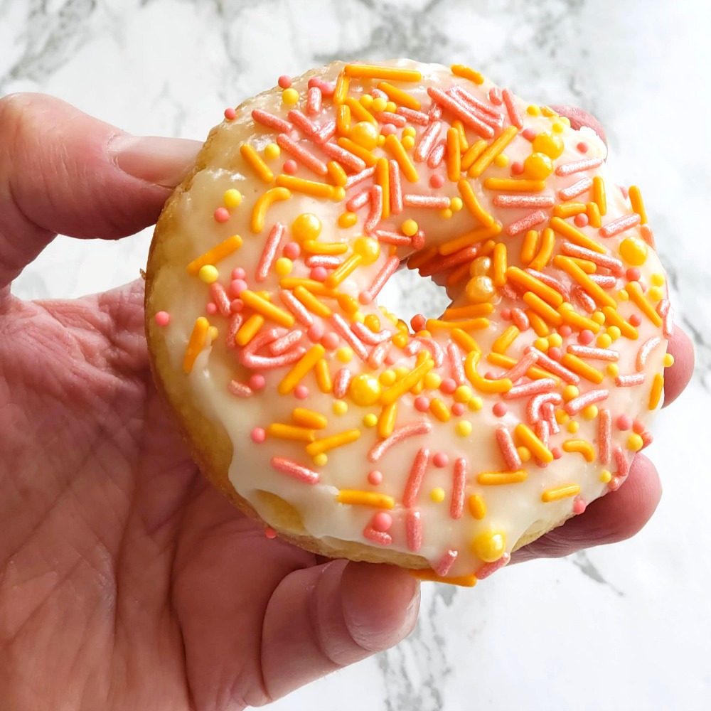 A hand holds a single Glazed Baked Donut with pink, orange and yellow sprinkles