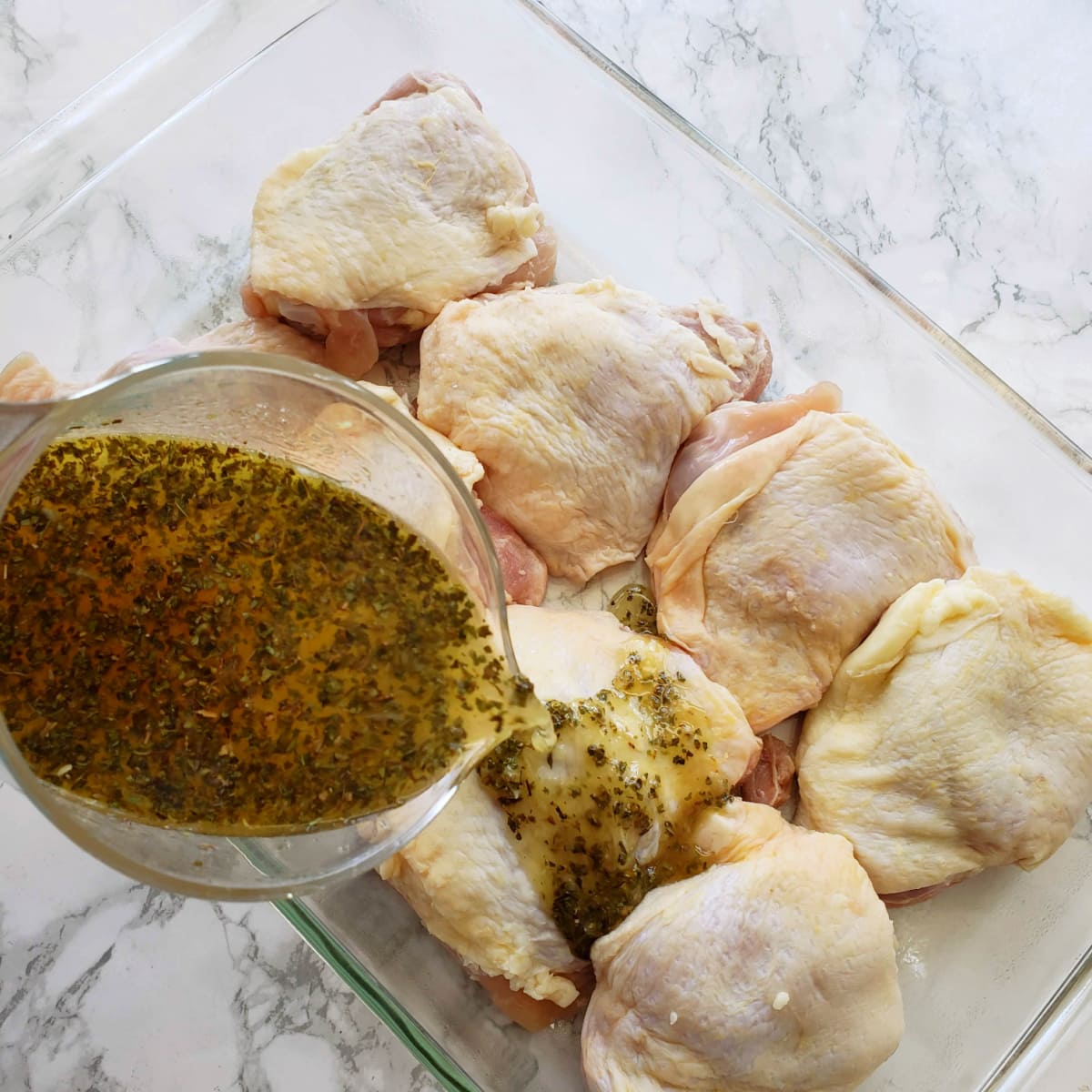 8 chicken thighs in a baking dish with marinade pouring over, set on a white marble counter