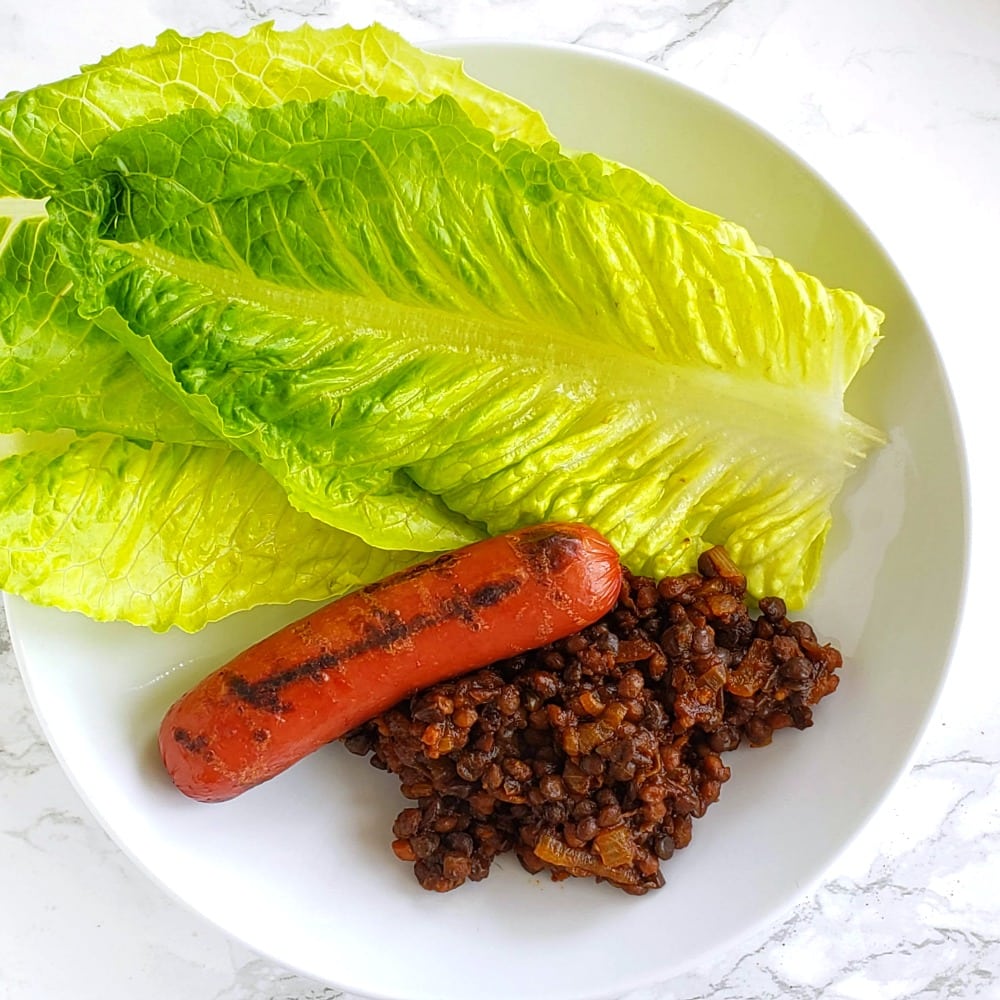 Barbecue Baked Lentils with a grilled hot dog and 3 spears Romaine lettuce in a white bowl