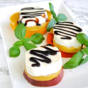 Slice of tomato topped with slice of polenta and slice of fresh Mozzarella, with a squiggle of balsamic glaze on top and basil alongside.