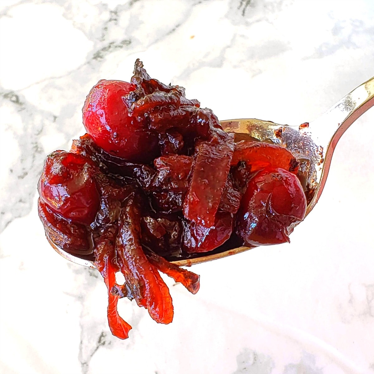 Cranberry Sauce with Caramelized Onions piled on a silver spoon with onions dangling against a white marble background