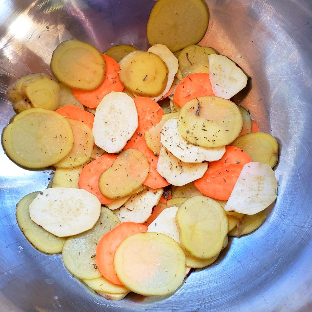 Potatoes layered in a big stainless steel mixing bowl with herbs