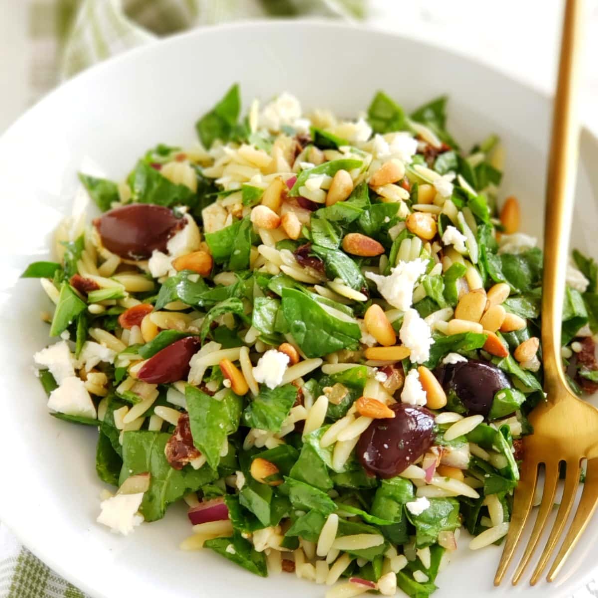 White bowl of Spinach Orzo Salad with a gold fork in the bowl and a green plaid tea towel underneath