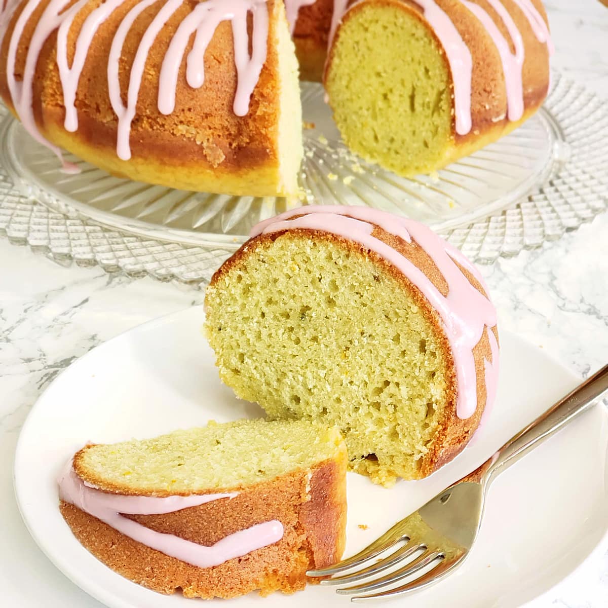 2 slices of Italian Blood Orange Olive Oil Bundt Cake on a white plate with fork, with the cake behind on a glass cake plate