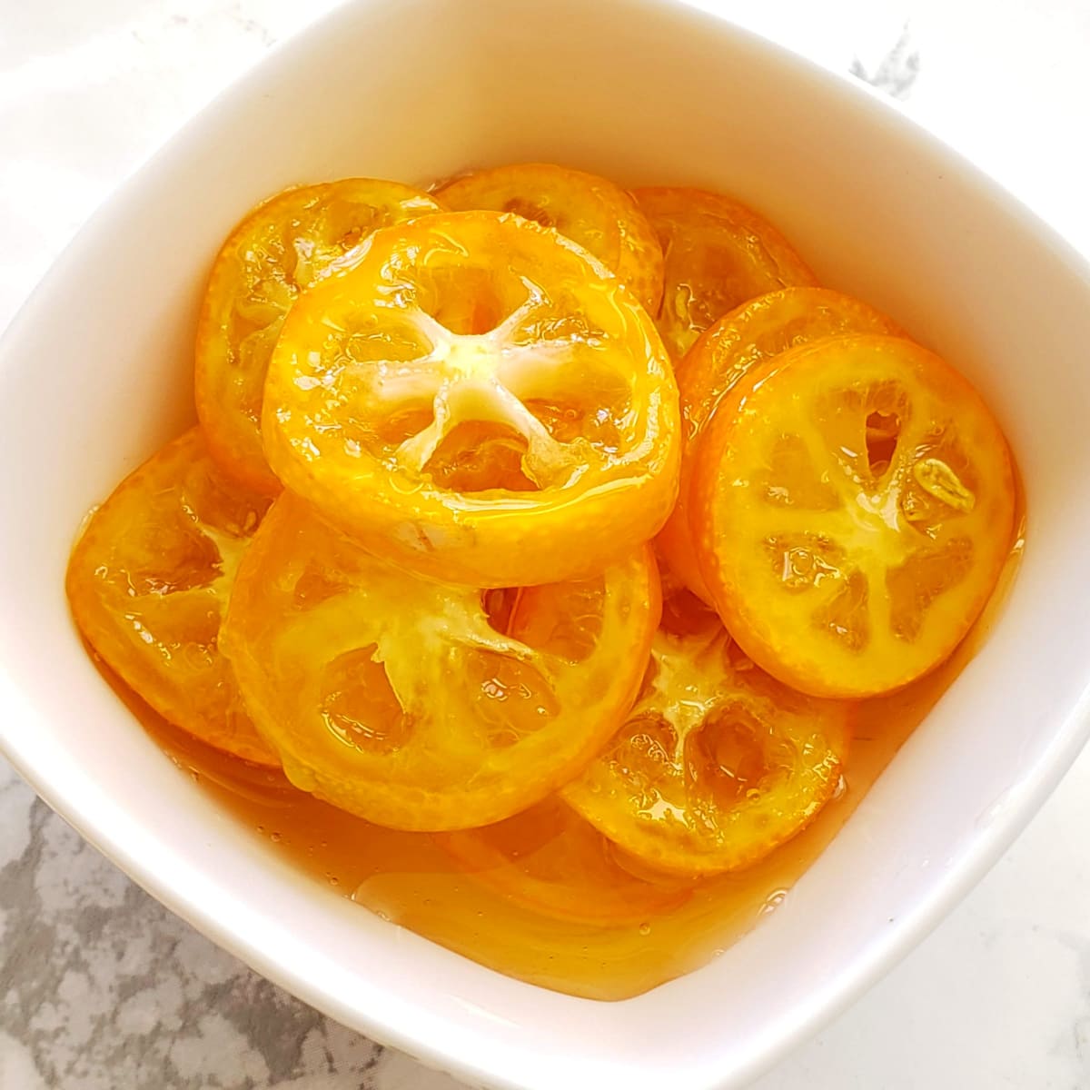 No-Cook Candied Kumquats slices in a white dish on a white marble counter on ShockinglyDelicious.com