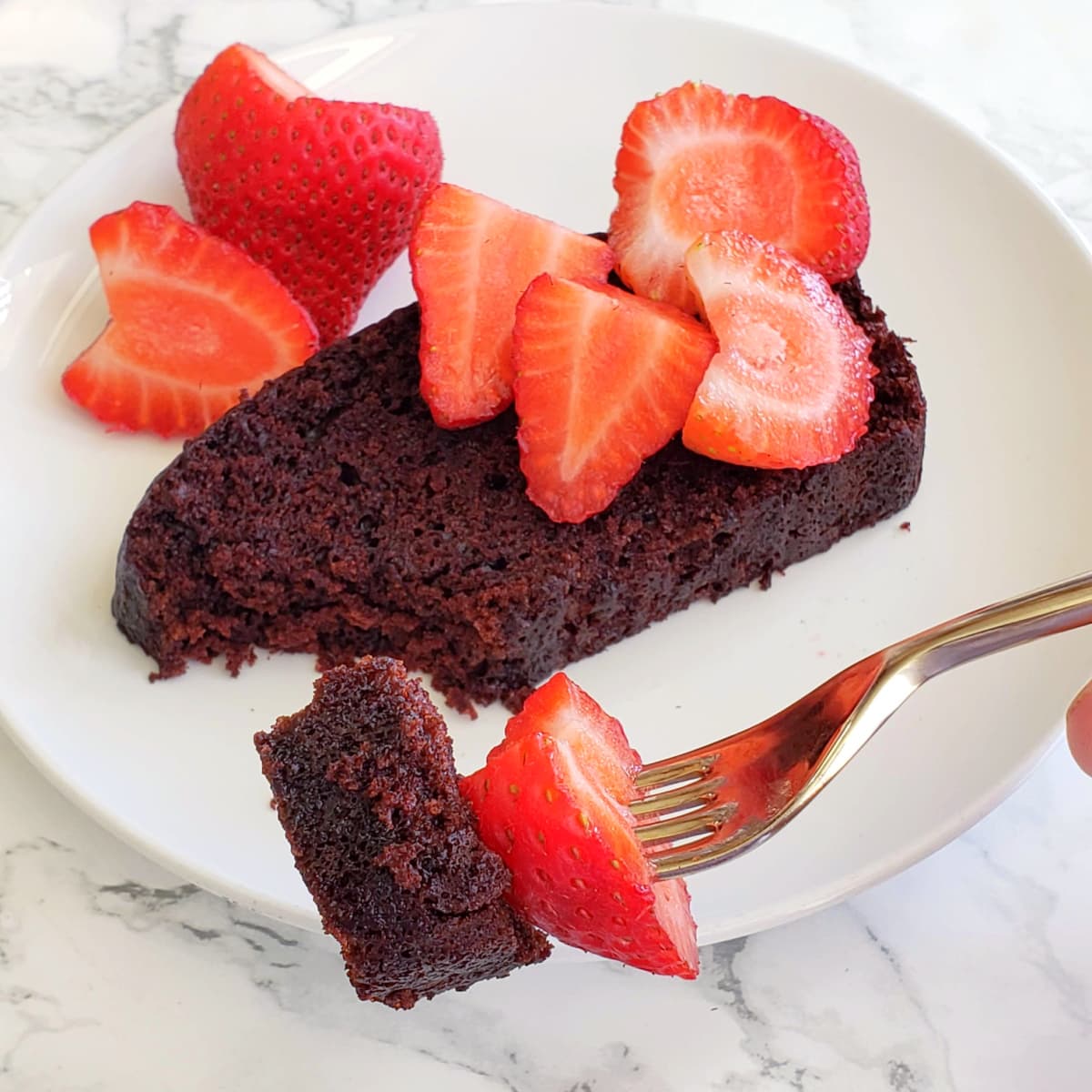 Slice of chocolate cake with strawberries on top, and a fork holding a bite of cake with a strawberry