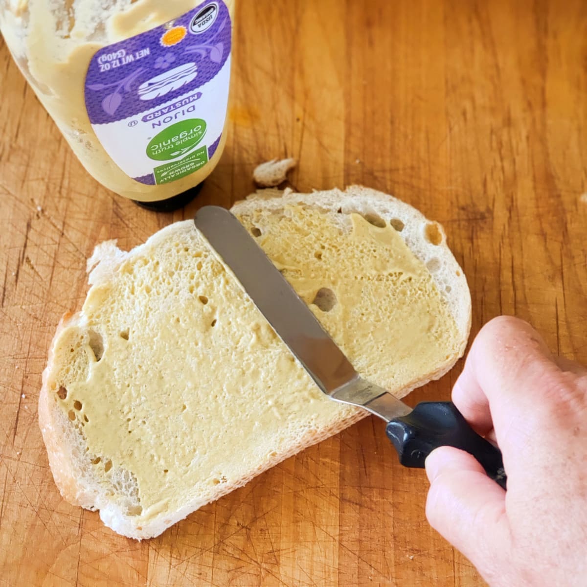 Spreading Dijon mustard on a sliced of sourdough bread with an offset spatula