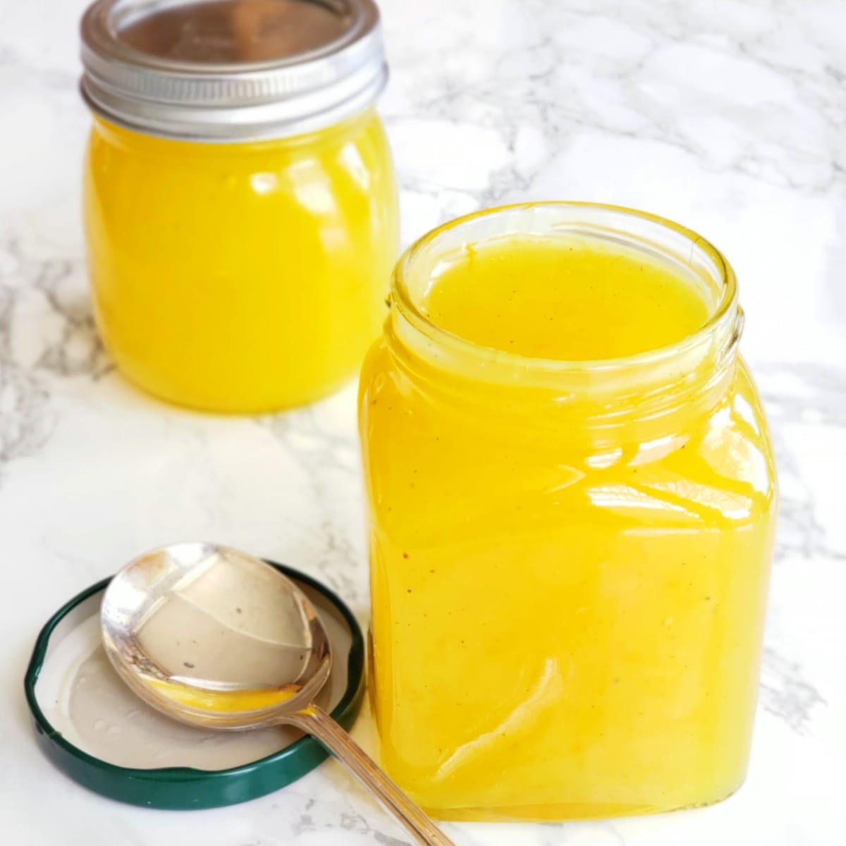 2 jars of lemon filling on a white marble counter. The jar in the foreground has its lid off and spoon resting in the lid