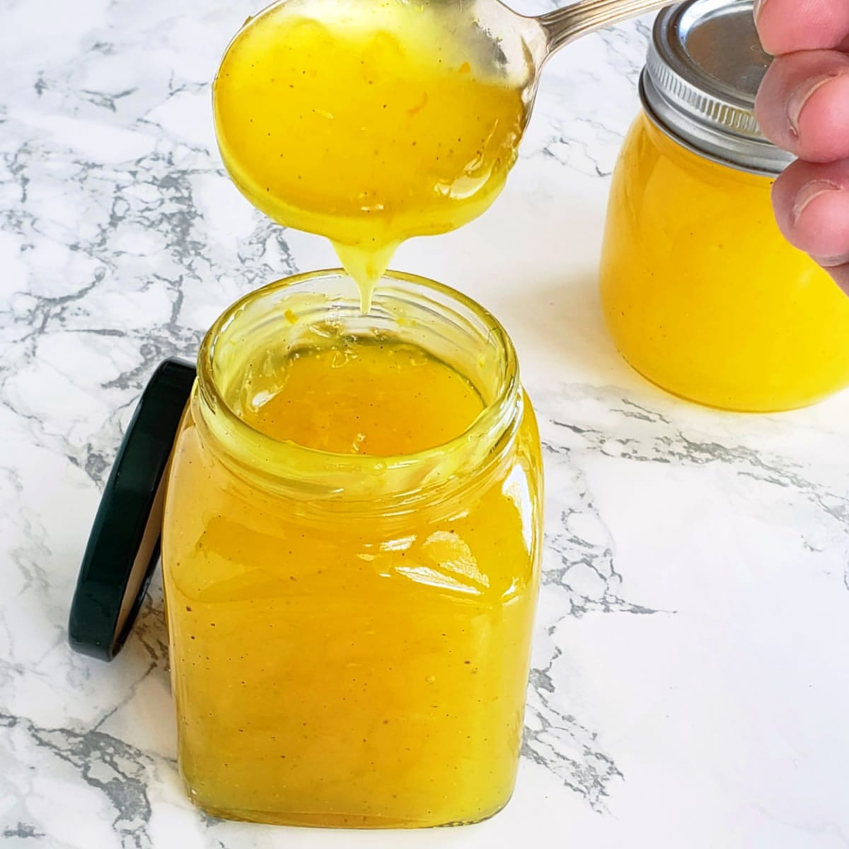 2 jars of lemon filling with a spoon dipping into the front jar, on a white marble counter
