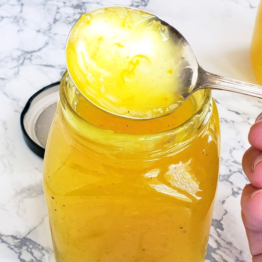 Silver spoon covered in lemon filling shows lemon zest, against a white marble counter background