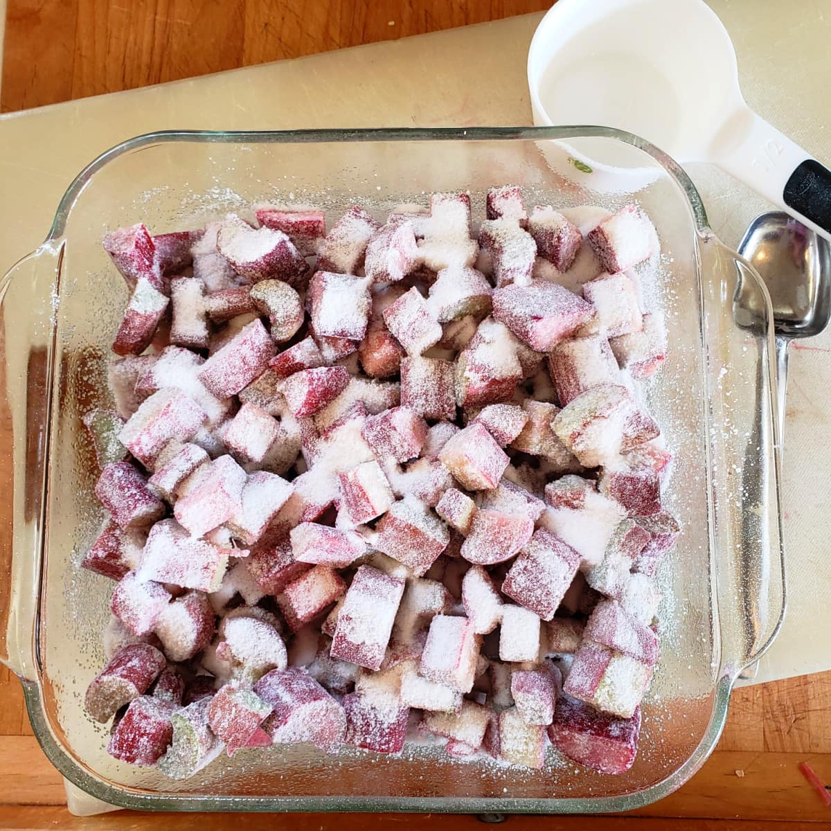 Rhubarb mixture in a square glass baking dish on a white cutting board with a white measuring cup and tablespoon measure beside it