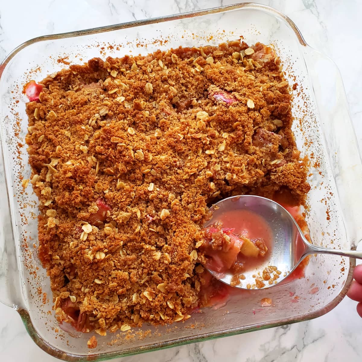 Rhubarb Crumble in a square glass baking dish with a large spoon and a corner of the crumble removed