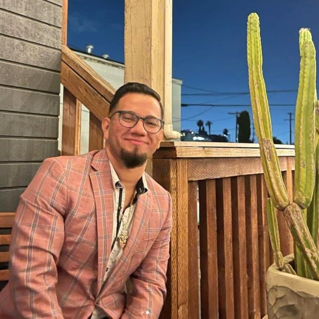 Chef Ronnie Munoz in a pink plaid suit sitting on a wooden porch at night