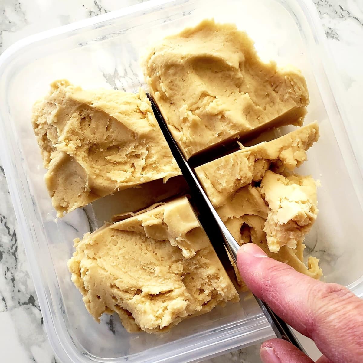 Dough in plastic container is being cut with a knife into 4 pieces, atop a white marble counter