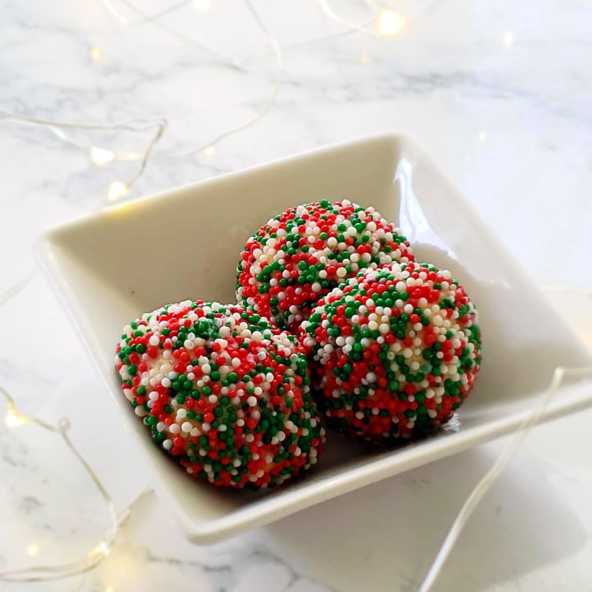 3 Cream Cheese Sprinkle Cookies in a white square bowl surrounded by twinkle lights on ShockinglyDelicious.com