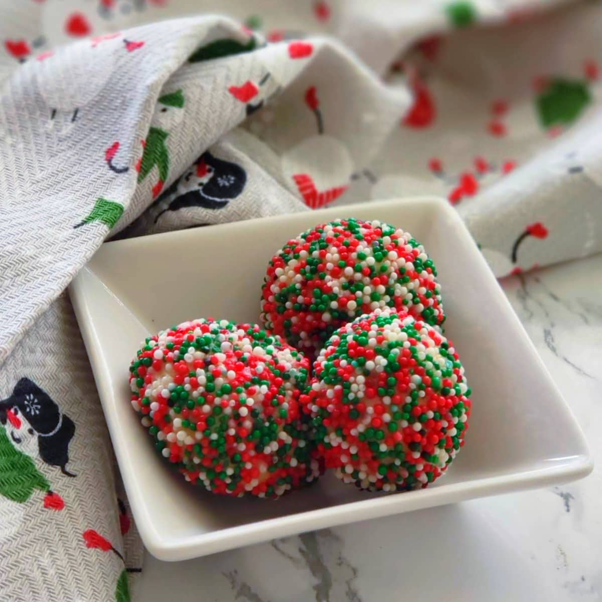 3 Cream Cheese Sprinkle Cookies in a square white bowl on a Christmas tea towel on ShockinglyDelicious.com