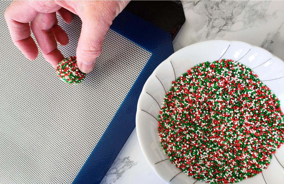 A hand puts cookie ball on a silicone lined baking sheet, with a white bowl of sprinkles alongside