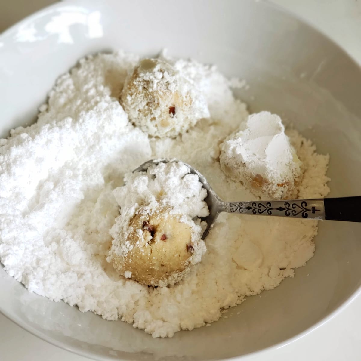 3 Mexican Wedding Cookies in a white bowl of powdered sugar with a spoon in the bowl