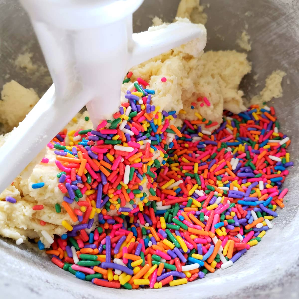 A pile of coloful sprinkles on top of cookie dough in a silver mixing bowl with a white beater
