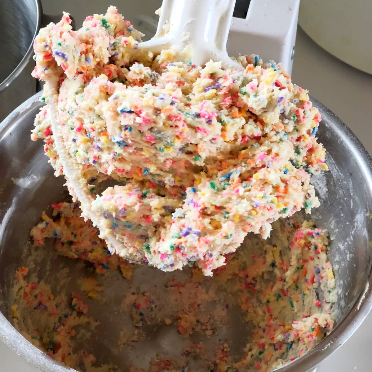 Thick dough with lots of sprinkles on a white beater and a silver mixing bowl for Confetti Sprinkle Cookies on ShockinglyDelicious.com