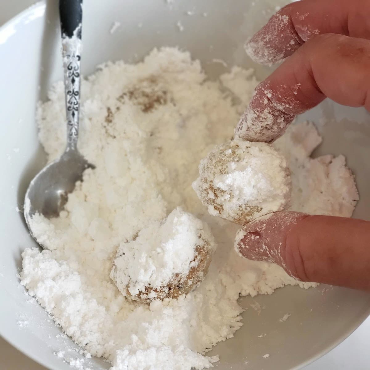 Fingers hold a Mexican Wedding Cookie in a bowl of powdered sugar with other cookies and a spoon also in the bowl