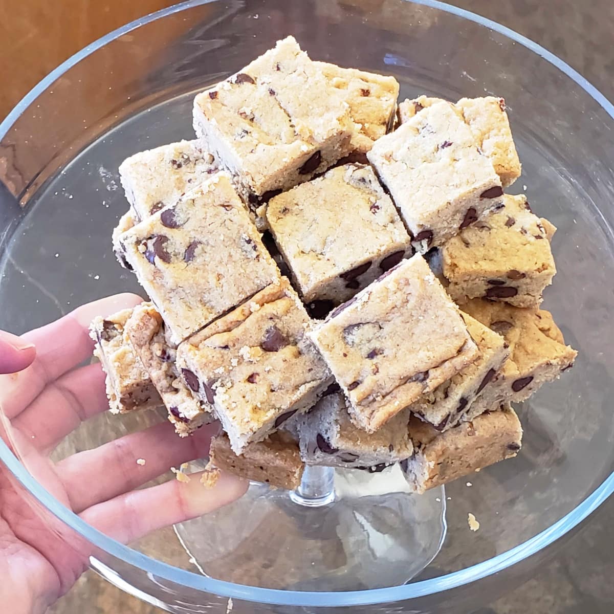 A hand holds a glass serving plate with Chocolate Chip Bar Cookies piled on the plate