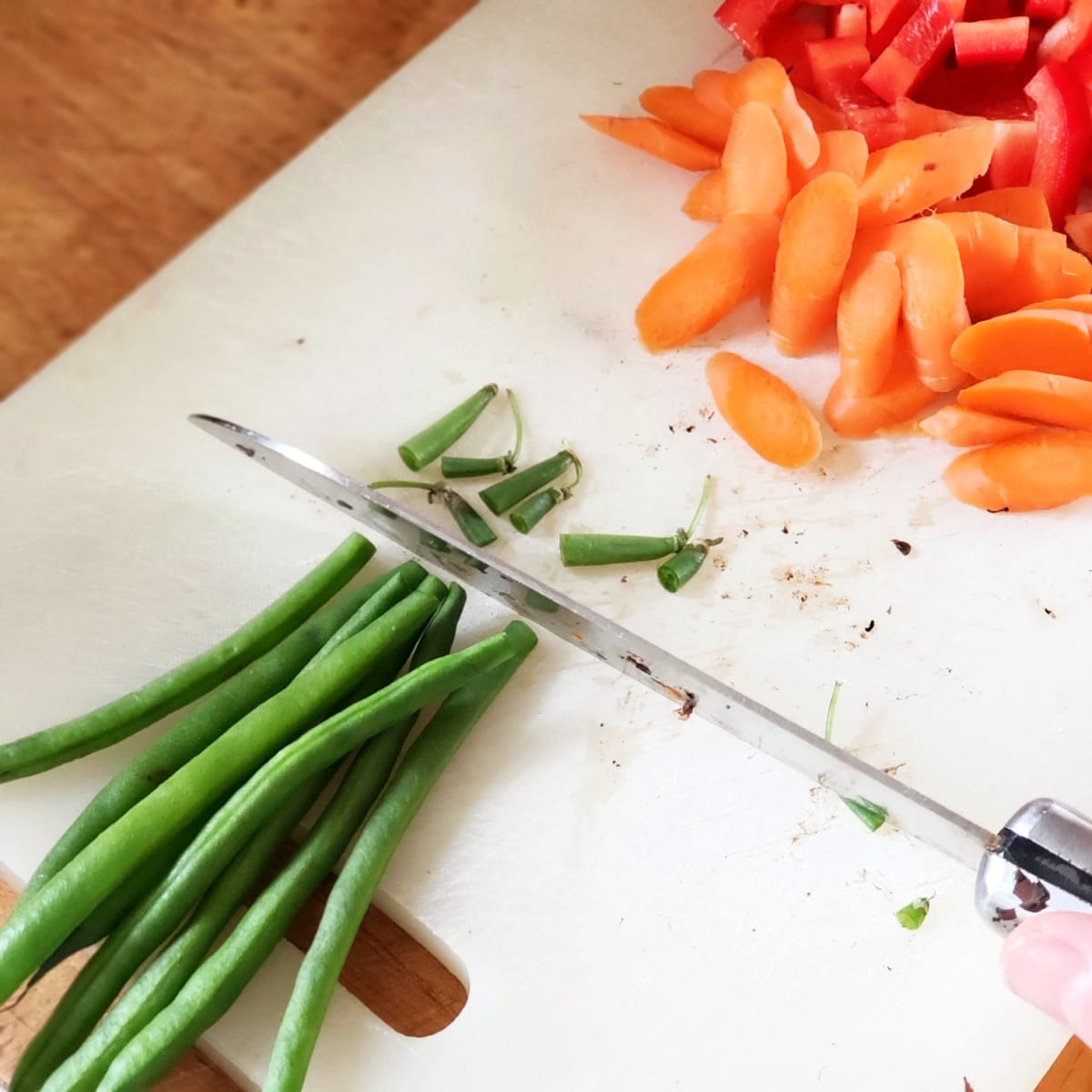 Chef's knife cuts green bean stems on a white cutting board, with sliced carrots to the upper left