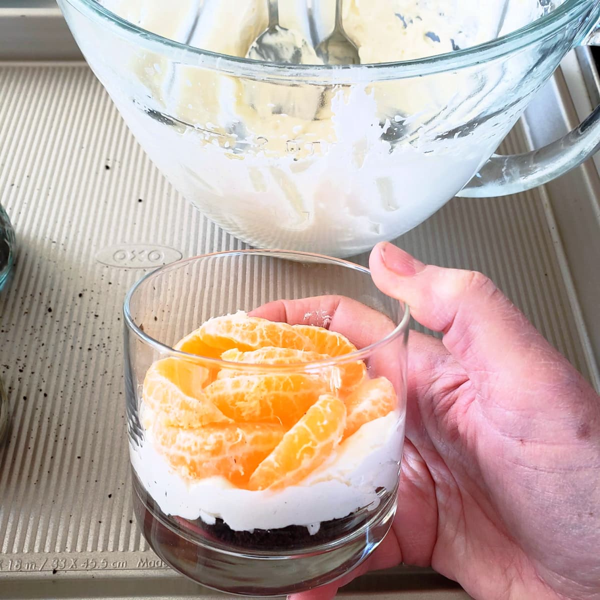 A hand holds a layered cheesecake dessert in a glass with a bowl full of white filling behind