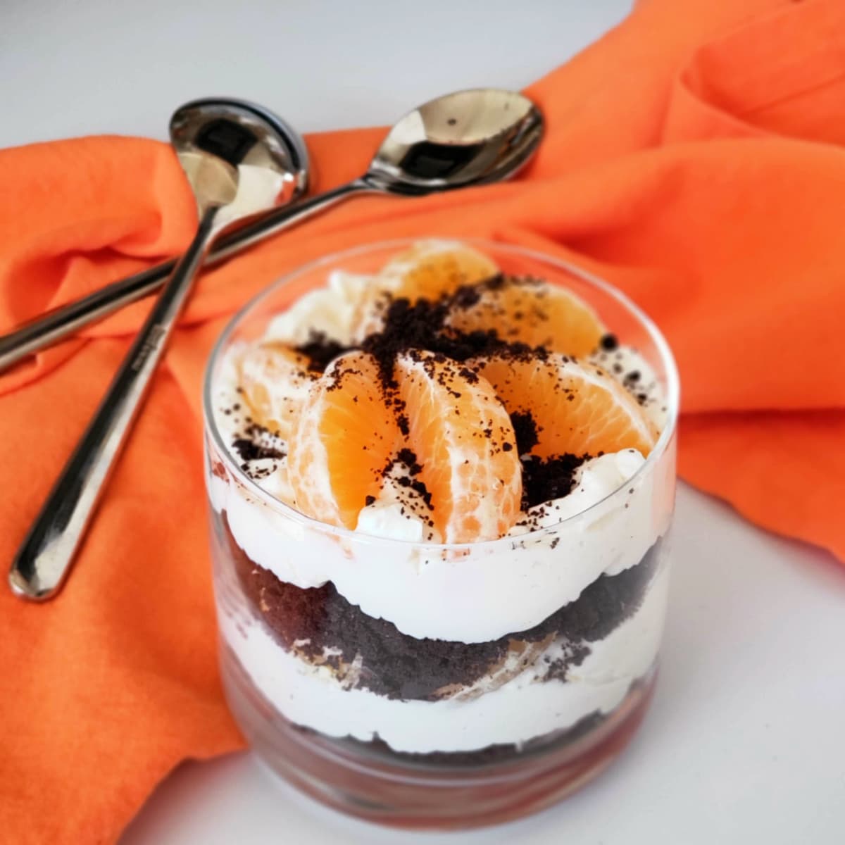 Layered cheesecake dessert with orange segments on top and spoons sitting on a bright orange napkin alongside