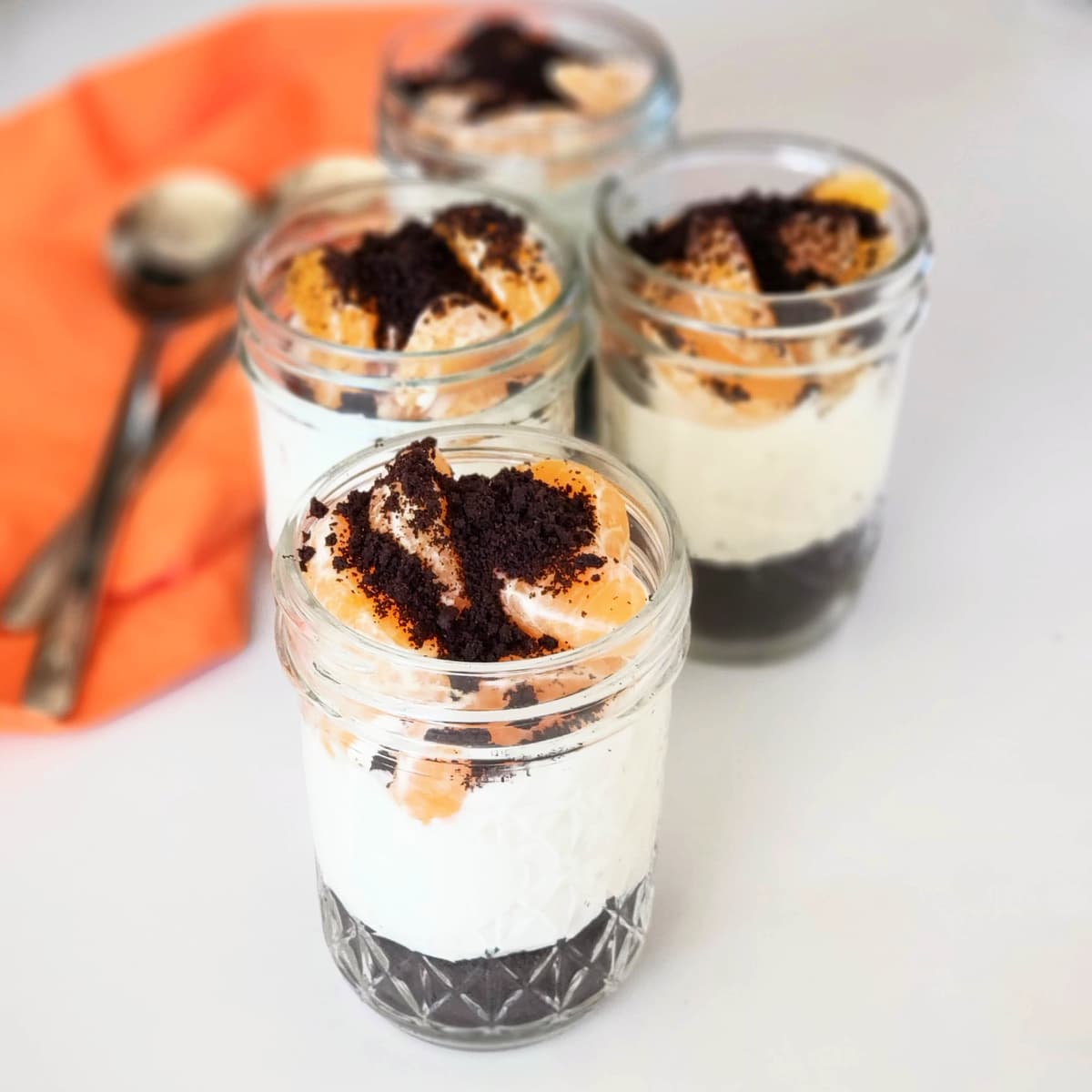 1 layered cheesecake dessert in the foreground and 3 in lesser focus in the background with an orange napkin and 2 spoons alongside