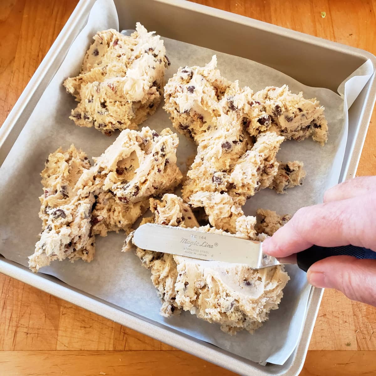Hand holding an offset spatula hovers over plops of thich dough in a gold toned baking pan
