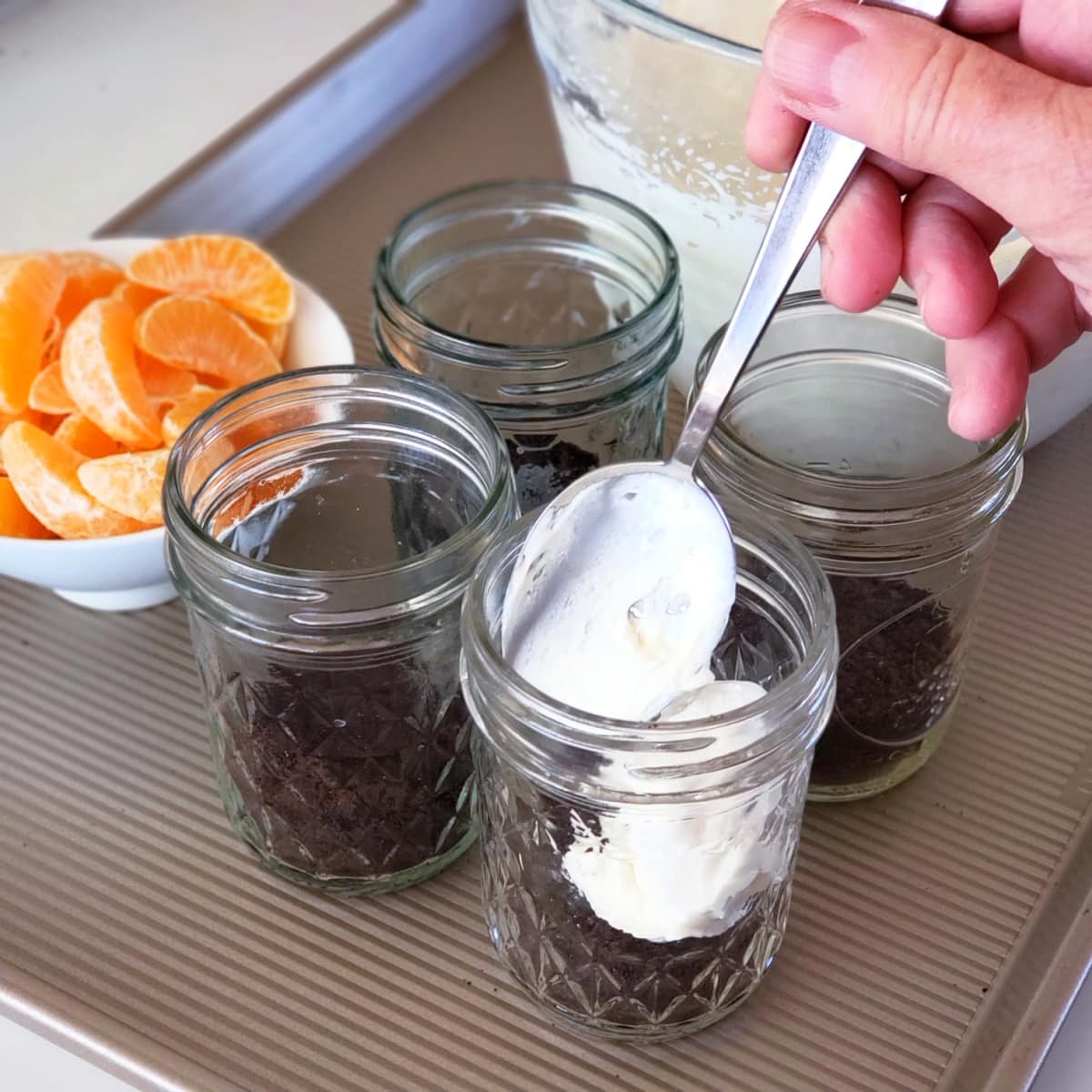 A hand holds a spoon that is dolloping cheesecake filling into jars that have chocolate cookie crumbs at the bottom. A bowl of tangerine segments is at the left.