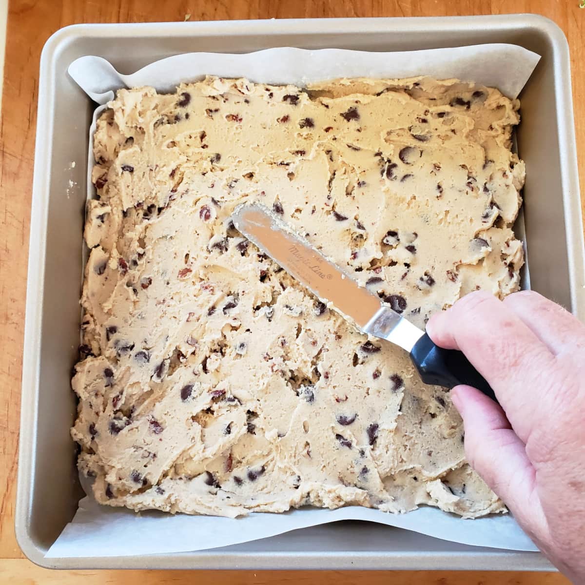 Hand holding an offset spatula spreads thick cookie dough in a gold toned baking pan