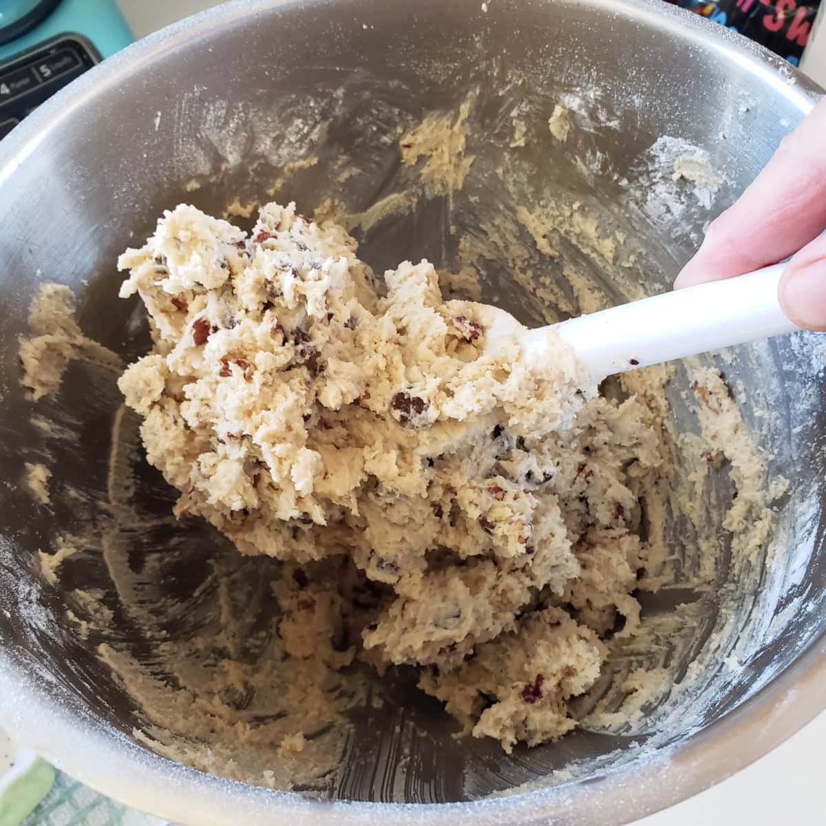Hand holding a white spoon stirs chocolate chips and pecans into the dough