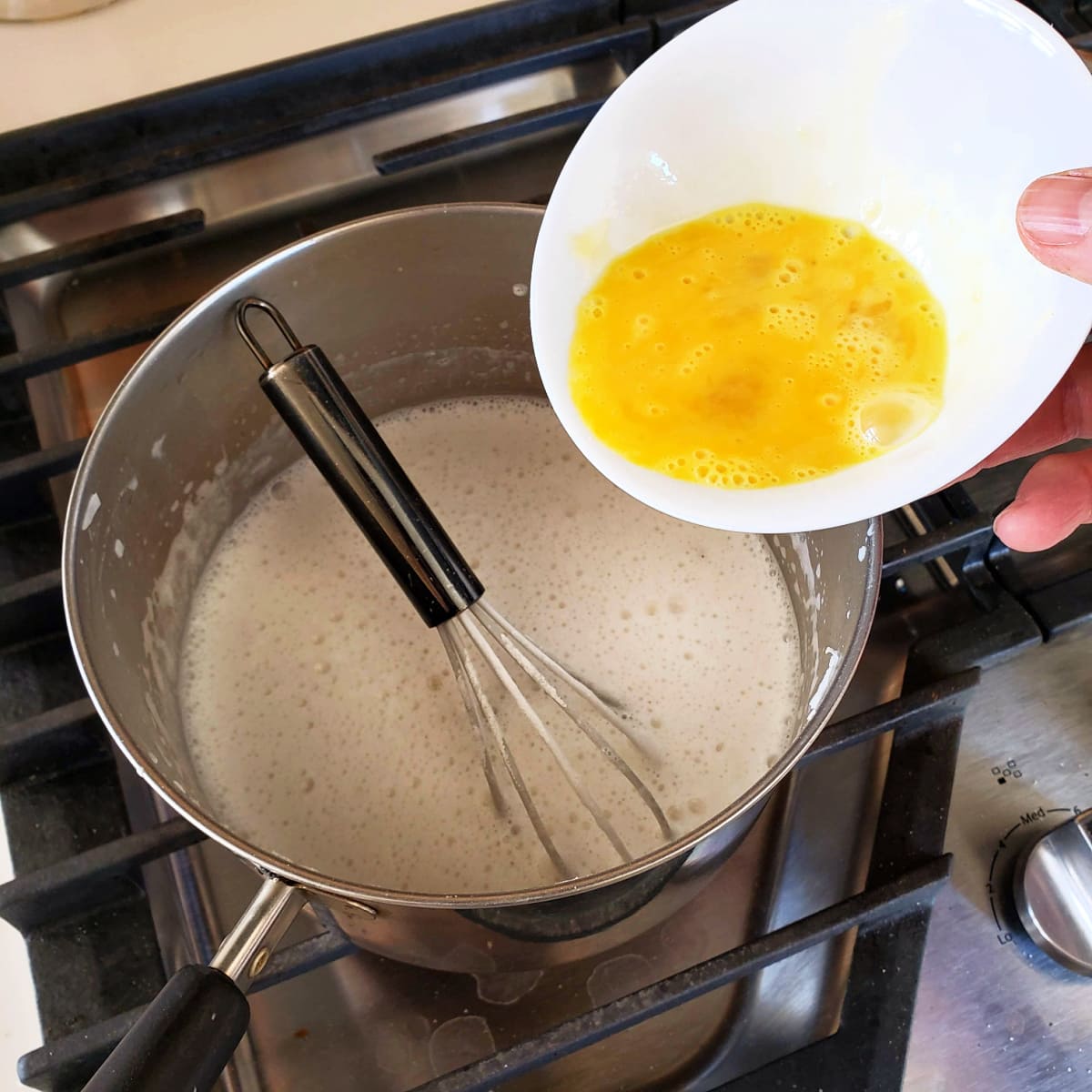 An egg in a small white bowl is being poured into a pan with milk and a whisk in it